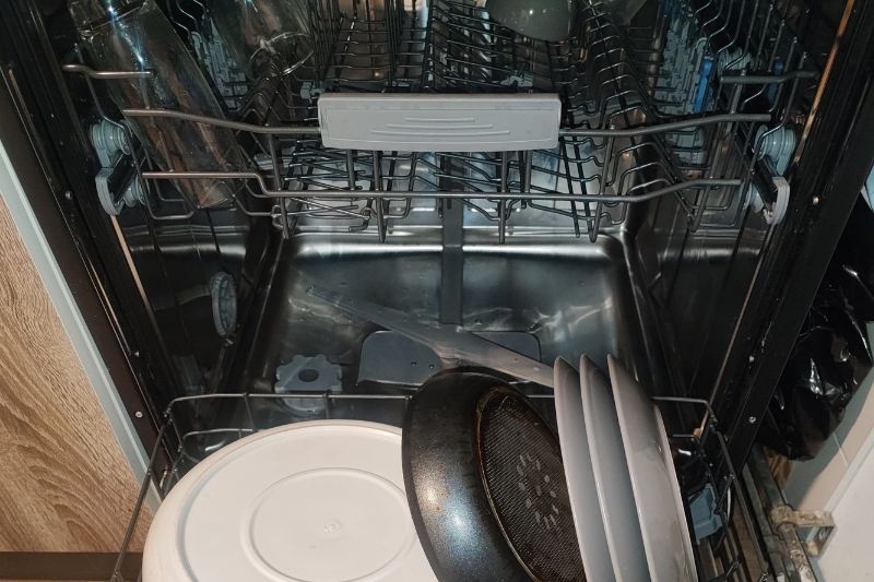 How to Clean a Dishwasher (Filter, Spray Arms & More) Open dishwasher with neatly loaded dishes, including a frying pan and plates on the lower rack. The stainless interior is clean and shiny.