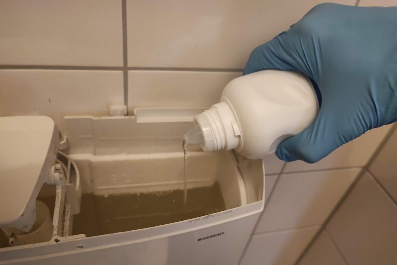 How to Clean a Toilet Tank (Step-by-Step Guide) Pouring white vinegar into an open toilet tank while wearing rubber gloves