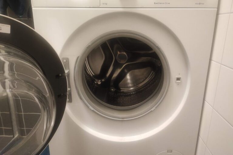 How to Clean a Front Load Washing Machine Step by Step Front-loading washing machine with open door, revealing an empty stainless steel drum. The surrounding area is tiled, giving a clean, modern appearance.