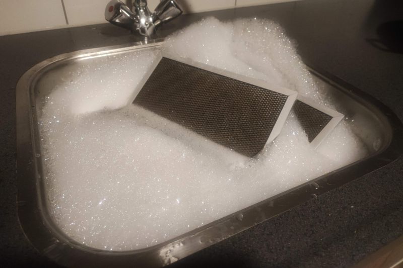 How to Clean a Range Hood Filter (Degrease in Minutes) Range hood filter soaking in hot soapy water in a kitchen sink