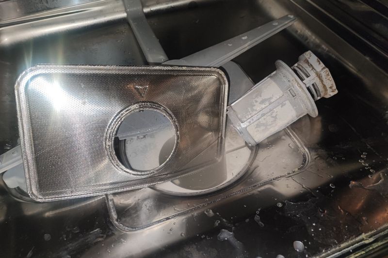 How to Clean a Dishwasher (Filter, Spray Arms & More) Removed dishwasher filter and spray arm on stainless steel sink, wet with water droplets; conveys maintenance or cleaning task.