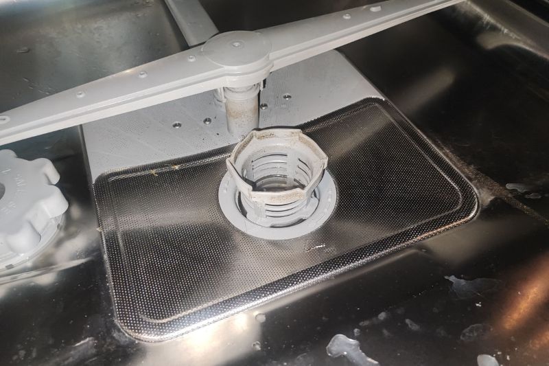 How to Clean a Dishwasher (Filter, Spray Arms & More) Close-up of a dishwasher interior showing the filter and spray arm. The metal surface appears slightly wet and filthy, with a worn filter indicating use.