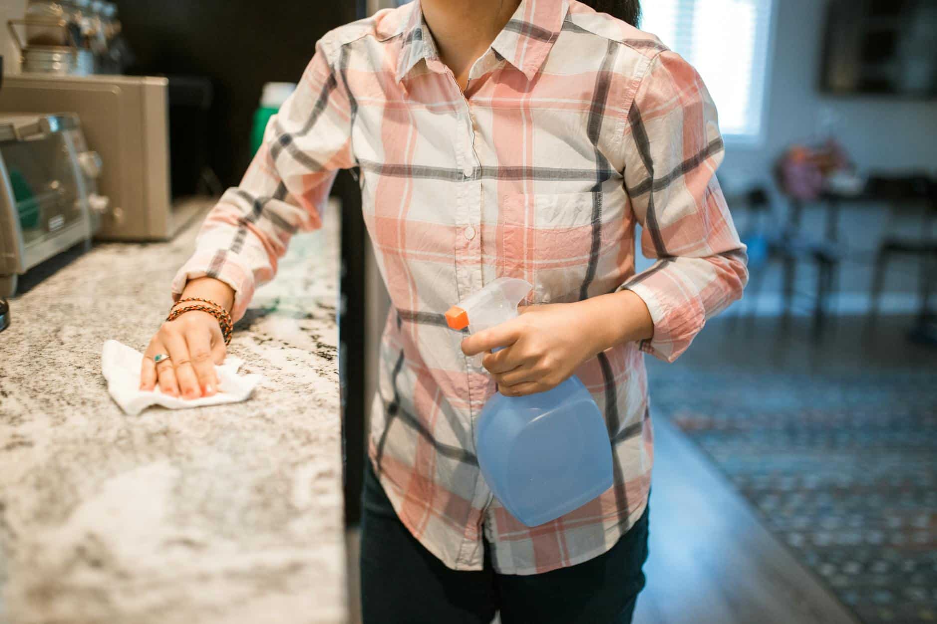 How to Deep Clean Your Kitchen: Full Sequence (2026) A person wiping a light granite countertop with a microfiber cloth and a spray bottle of gentle cleaner