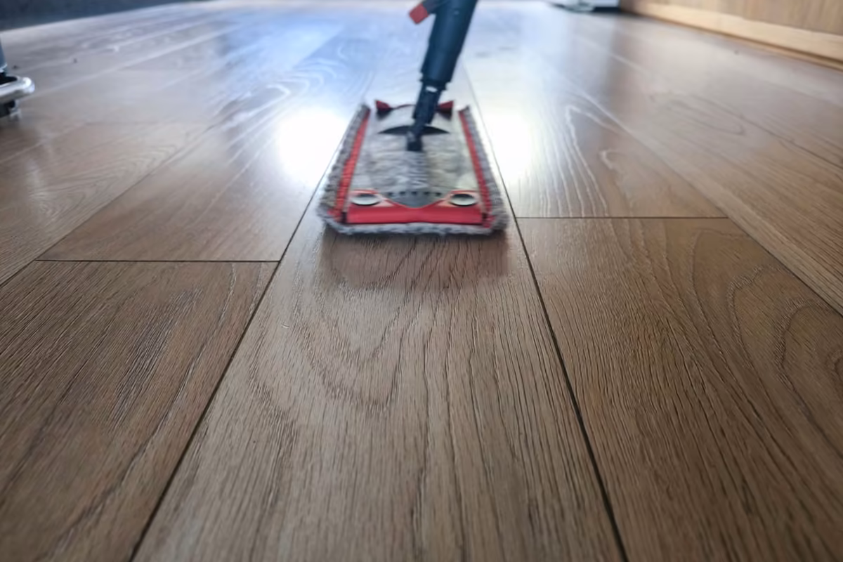 Low angle view of a flat microfiber mop with a red frame cleaning a wood-look plank floor, light reflecting off the boards.