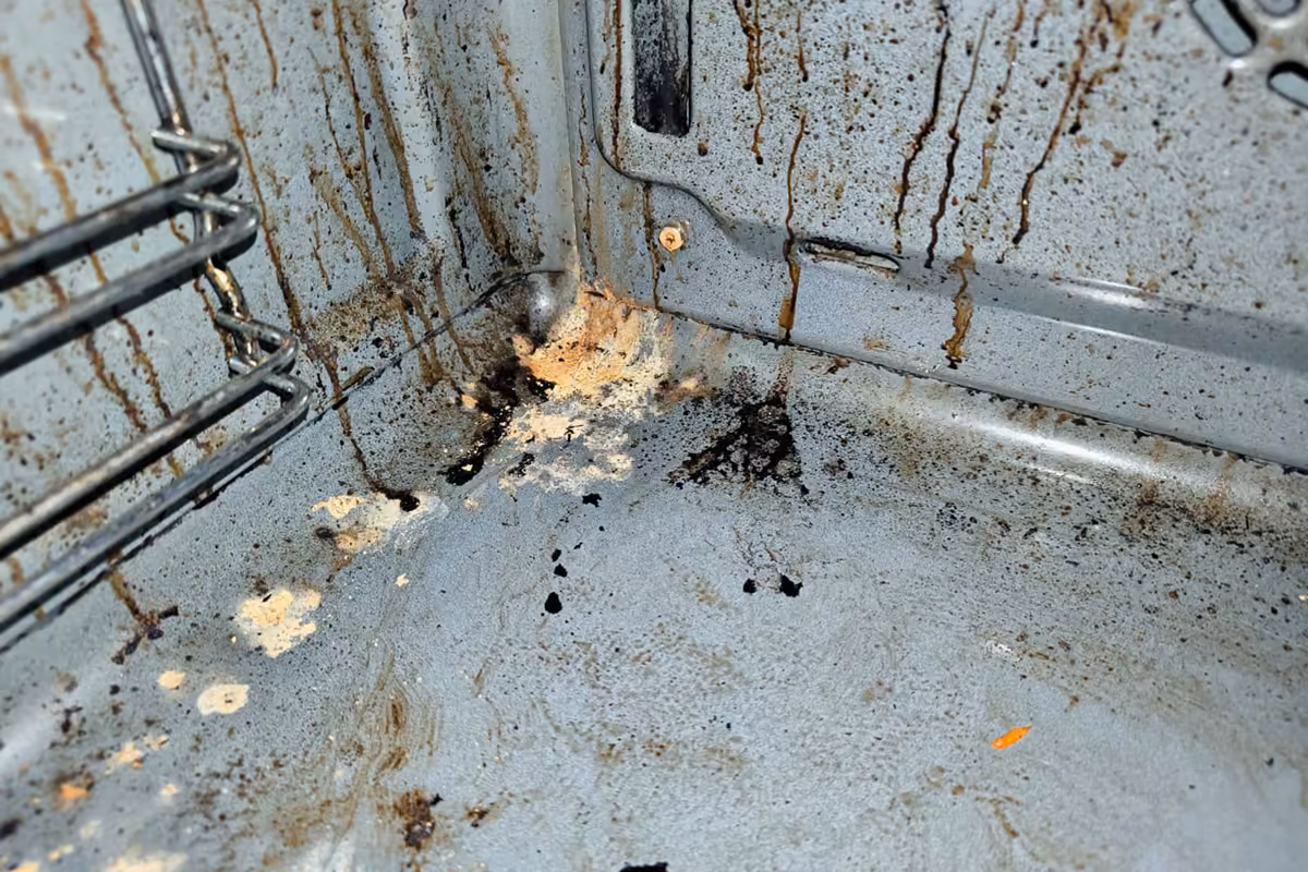 Inside of a dirty oven with brown grease drips running down the back wall, burnt black spots and crusted food residue on the oven floor, and the rack supports on the left.
