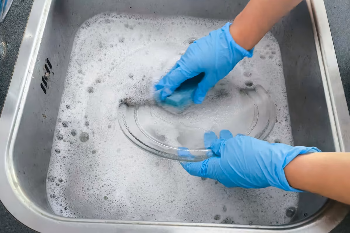 Two gloved hands washing a glass microwave turntable plate with a sponge in a stainless steel sink full of soapy water.