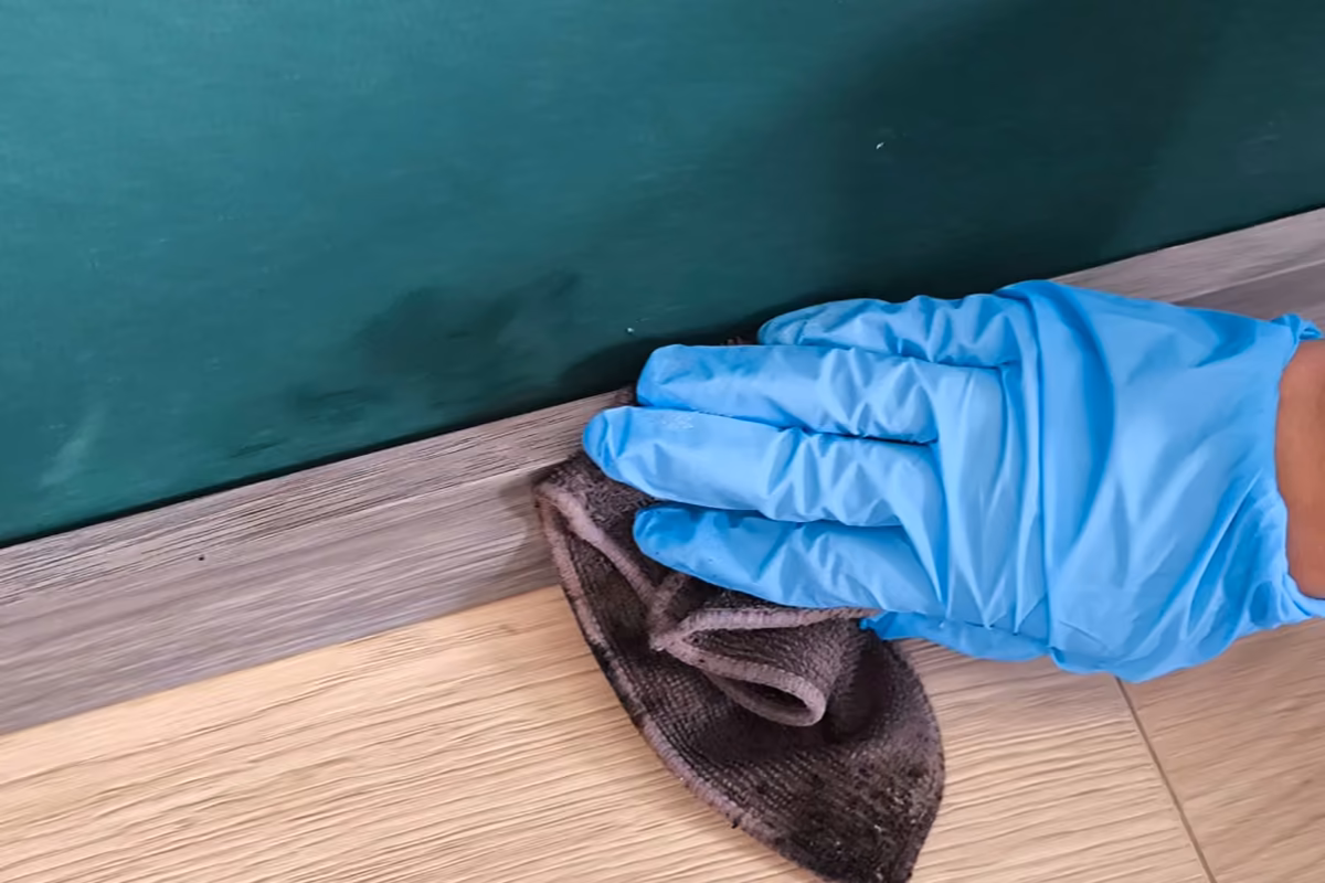 Gloved hand wiping a grey baseboard with a microfiber cloth that is black with dust, against a dark green wall and a light wood floor.